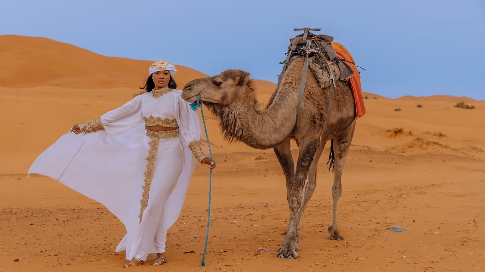 Mujer con caftán blanco junto a un camello en las dunas de Erg Chebbi